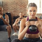 Group of fit people holding kettle bell during squatting exercise at cross training gym. Fitness girl and men lifting kettlebell during strength training exercising. Group of young people doing squat with kettle bell.
